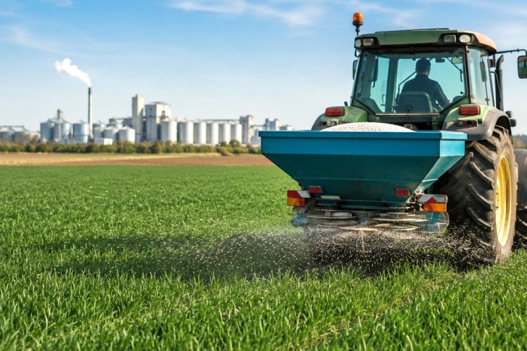 Landwirt bringt Dünger auf einem Feld aus, im Hintergrund Industrie- und Logistikanlagen als Symbol für globale Lieferketten.