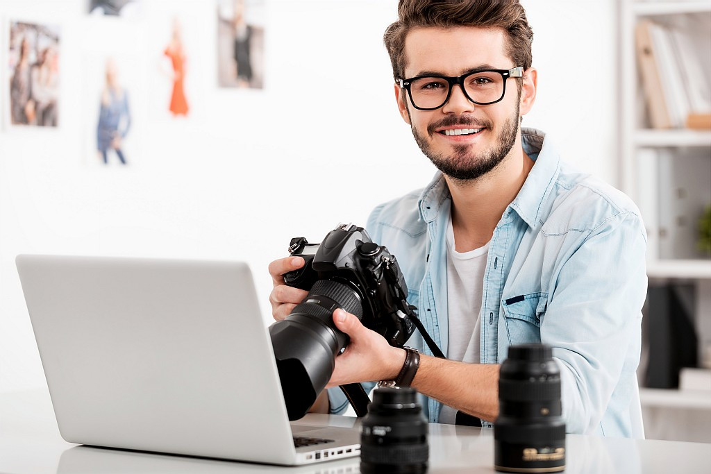 Glücklicher junger Fotograf, sitzt vor einem Laptop und hält eine Kamera